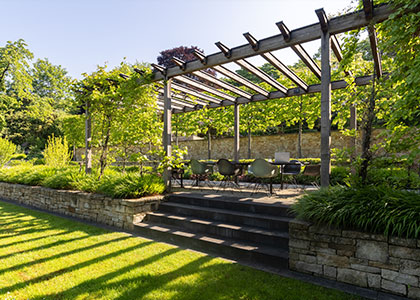 Begrünte Pergola mit Sitzbereich, Stufen und Natursteinmauern im Garten.