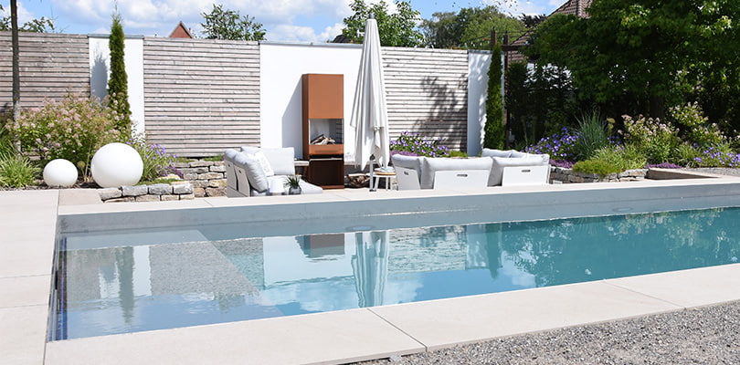 Moderner Naturpool mit klarem Wasser, Loungebereich und Feuerstelle im Garten.